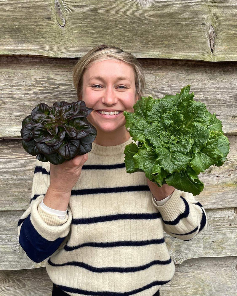 Woman smiling holding two lettuce plants in front of a wooden cabin wall